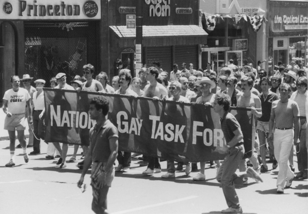 A black and white photo of people marching in the street behind a National Gay Task Force banner.