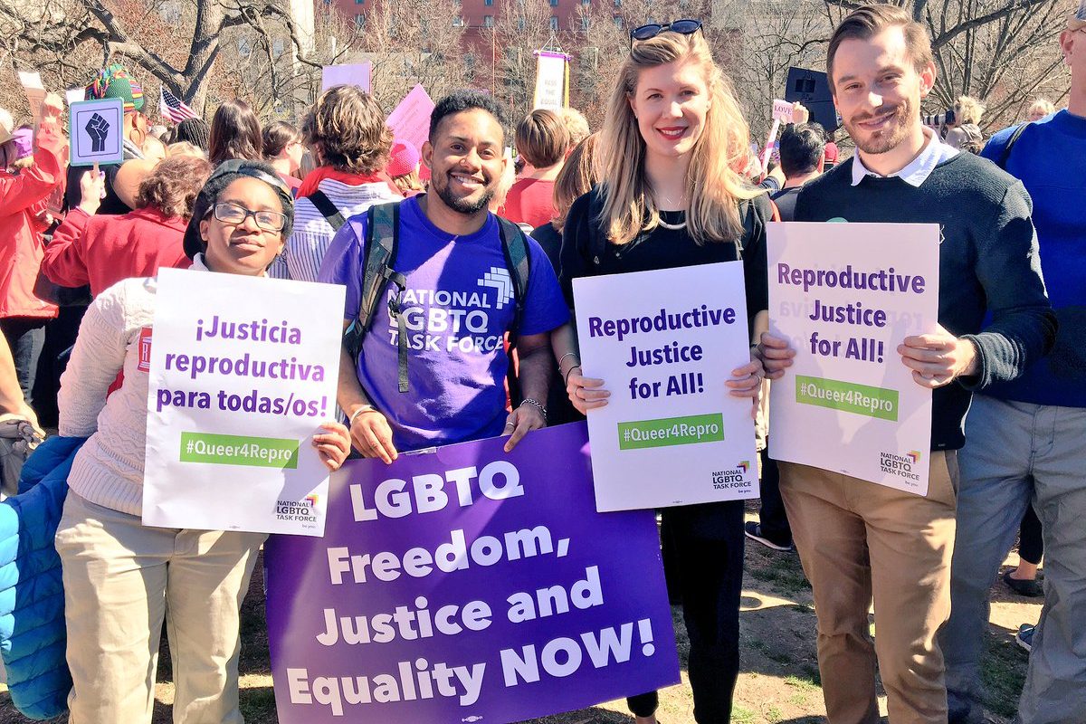 A group of young black and white advocates hold signs at an outdoor gathering. Their signs read "Justicia reproductive para todas/os," "LGBTQ Freedom, Justice and Equality NOW," and "Reproductive Justice for All." The signs include the hashtag #Queer4Repro. All four people are smiling and facing the sun.