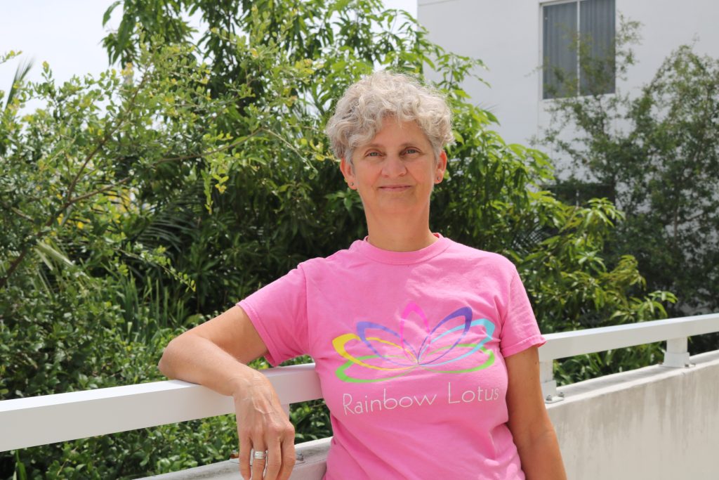 The image features Mx. Contance Collins, JD, the "2023 Eddy McIntyre Community Service Award" recipient. Collins has short, curly hair and is standing in front of trees wearing a pink t-shirt. Collins' t-shirt catches attention as it features a lotus flower design. The lotus flower design is emphasized as the text "Rainbow Lotus" is displayed just below, providing further context to the flower design.